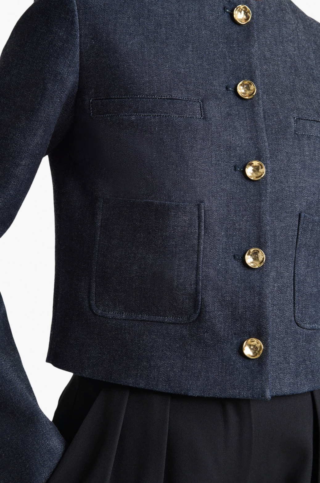 A close-up of the Altuzarra Bernadette Jacket, an Italian-made dark blue denim piece with gold buttons and patch pockets, styled over black pants.