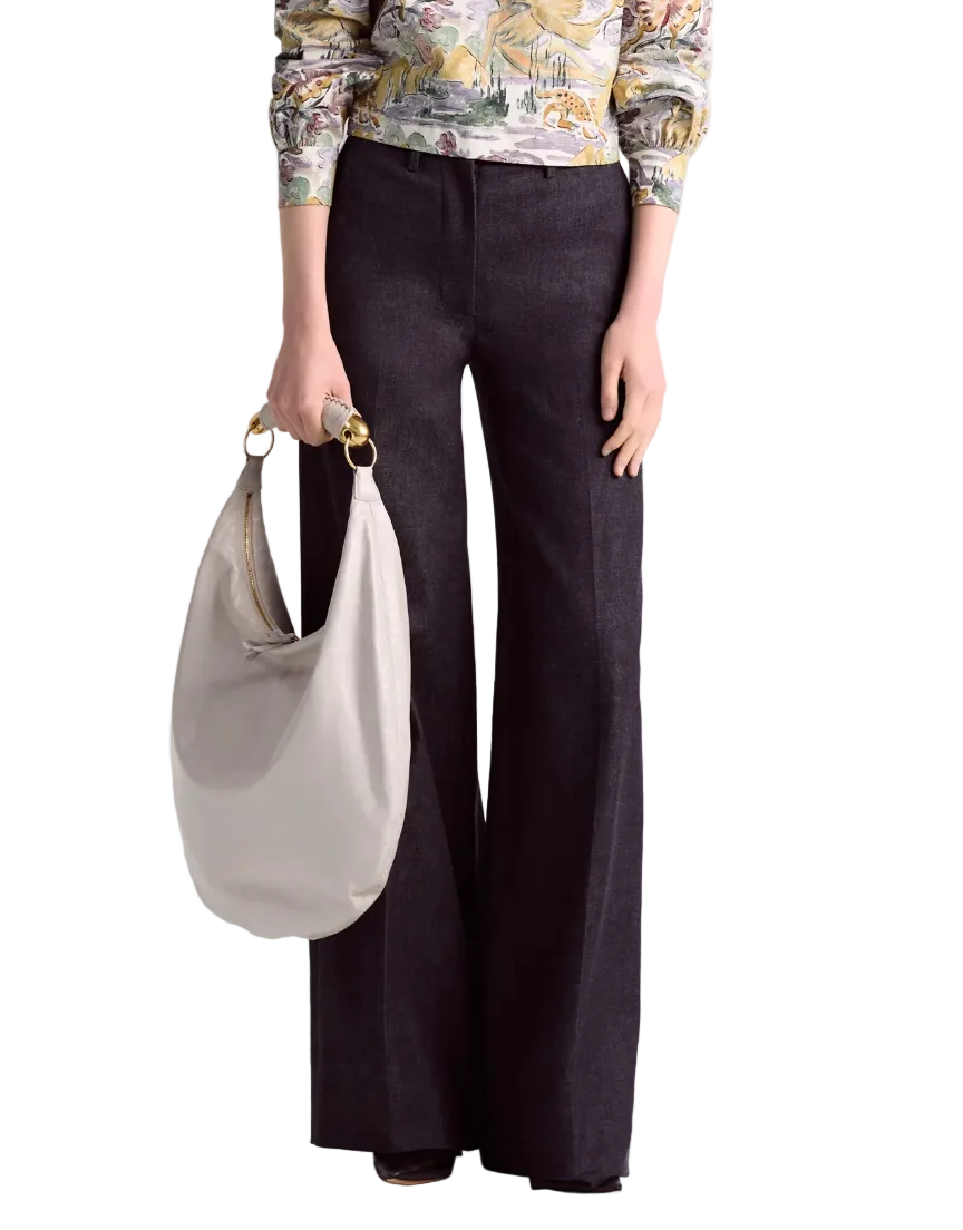 A person wearing the Altuzarra Rudy Pant, a high-waisted wide-leg black style, paired with a patterned long-sleeve top and carrying a large light beige handbag.