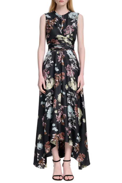 A woman stands with arms at her sides in the Altuzarra Penny Dress, a sleeveless, high-low floral dress with a colorful print, styled with black strappy heels—perfect for wedding guests.