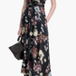A woman wears the Altuzarra Penny Dress—a sleeveless, floor-length black floral gown with an asymmetrical hem—styled for a wedding with a black handbag and high-heeled sandals.