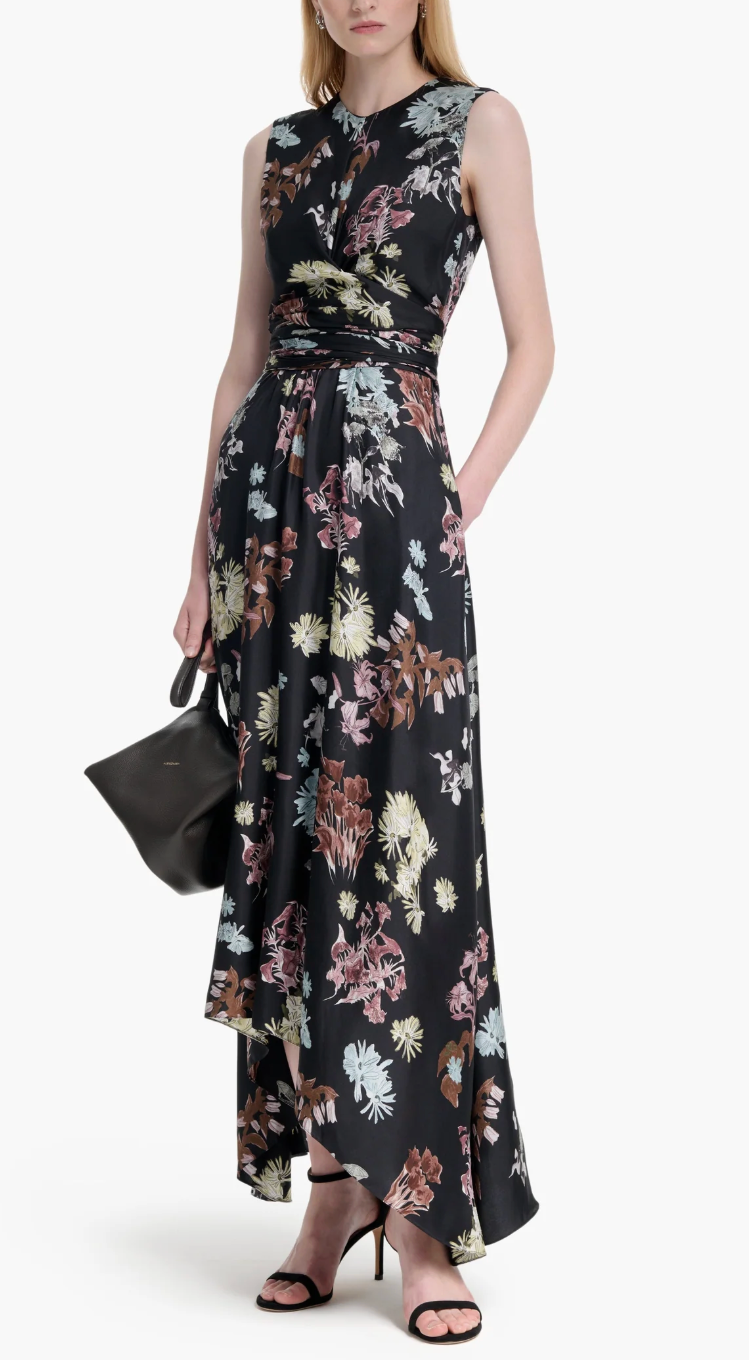 A woman wears the Altuzarra Penny Dress—a sleeveless, floor-length black floral gown with an asymmetrical hem—styled for a wedding with a black handbag and high-heeled sandals.