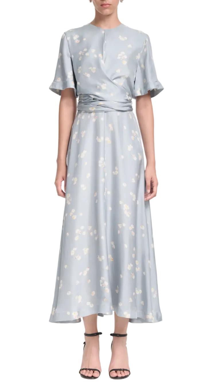 A woman wears the Altuzarra Kirsty Dress, a light blue ankle-length floral piece with a cinched waist and wrap bodice, paired with black strappy heels.