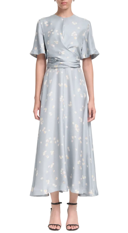 A woman wears the Altuzarra Kirsty Dress, a light blue ankle-length floral piece with a cinched waist and wrap bodice, paired with black strappy heels.