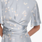 A person wears the Altuzarra Kirsty Dress—a light blue silk short-sleeve dress with a subtle floral print, wrap bodice, gathered waist, and high neckline. Their head is turned slightly to the side.