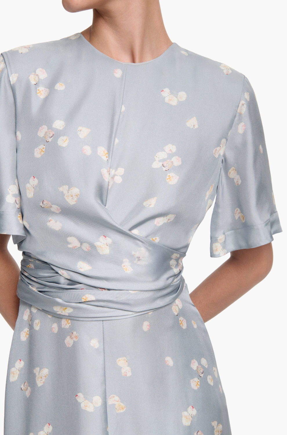 A person wears the Altuzarra Kirsty Dress—a light blue silk short-sleeve dress with a subtle floral print, wrap bodice, gathered waist, and high neckline. Their head is turned slightly to the side.