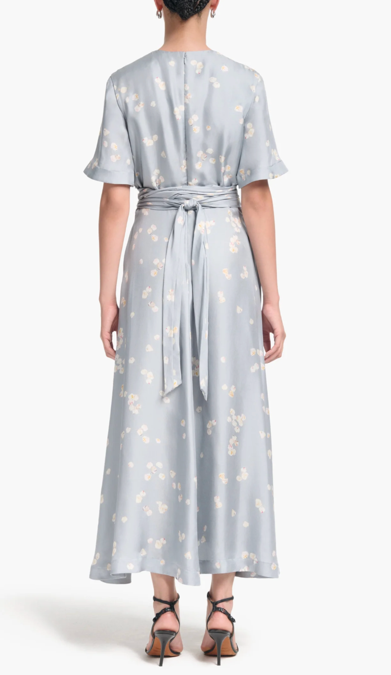 A person faces away, wearing the Altuzarra Kirsty Dress—a light blue floral short-sleeve wrap dress with waist tie and wrap bodice—paired with black heeled sandals.