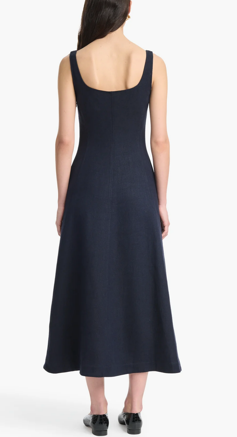 A woman faces away, wearing the sleeveless dark blue Altuzarra Orrie Dress with vintage-inspired buttons and black shoes, standing against a plain white background.