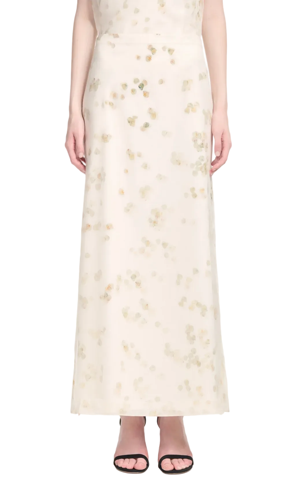 A person is shown from the shoulders down wearing the Altuzarra Linnie Skirt—a long white maxi with subtle petal designs—paired with black open-toe sandals.