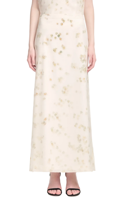 A person is shown from the shoulders down wearing the Altuzarra Linnie Skirt—a long white maxi with subtle petal designs—paired with black open-toe sandals.