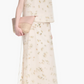 A woman wears the Altuzarra Linnie Skirt—a cream maxi with subtle petal motifs—paired with a matching sleeveless top, beige clutch, and black heeled sandals.
