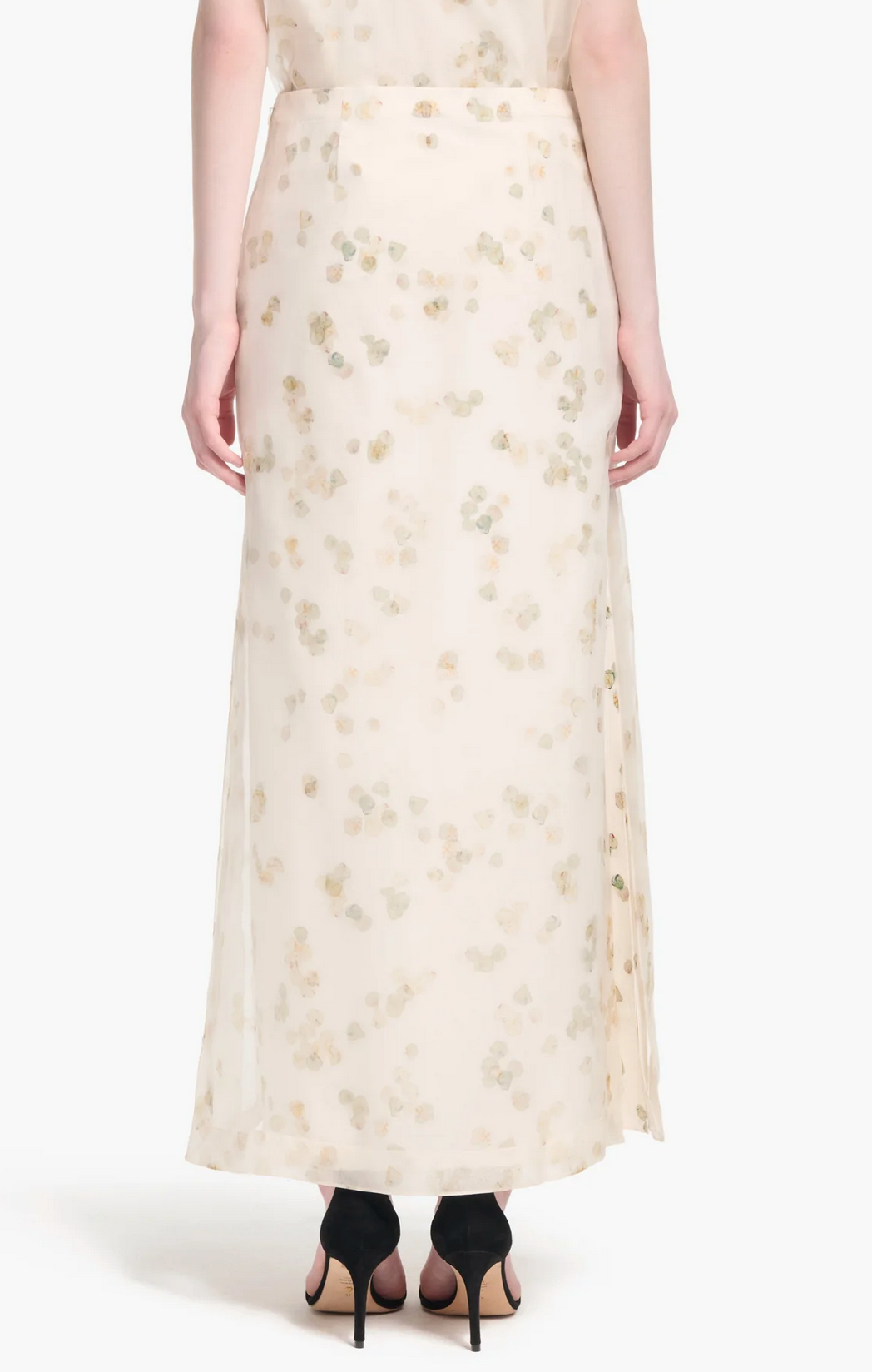 A woman in the Altuzarra Linnie Skirt—a long, sheer cream skirt with subtle petal motifs and a flowing maxi silhouette—paired with black high-heeled sandals, viewed from behind.