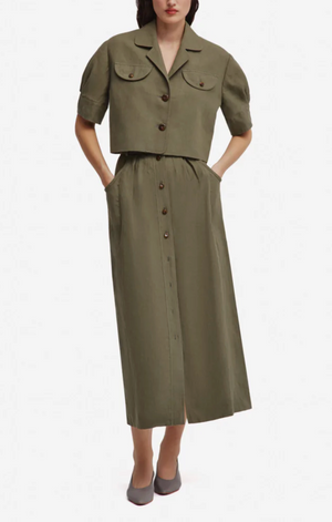 Wearing the Blaze Milano Midday Sun Nija Skirt in palm green linen, paired with a button-down, short-sleeve cropped jacket, hands in pockets and gray flats.