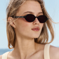 A woman with blonde hair wears the Elvie Sunglasses by Jimmy Fairly, which feature a sleek cellulose acetate frame in round black design, paired with a cream top as she stands outdoors beneath a blue sky.