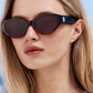 A woman with straight blonde hair and a neutral expression is wearing Jimmy Fairly’s Meli Icons sunglasses, which feature a chic cellulose acetate butterfly frame and dark oval lenses, set against a light blue background.