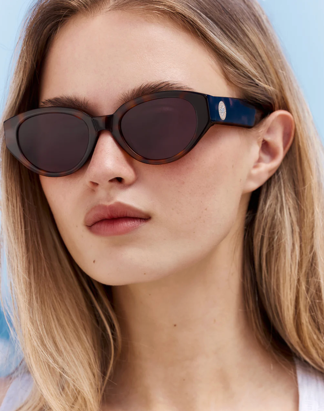 A woman with straight blonde hair and a neutral expression is wearing Jimmy Fairly’s Meli Icons sunglasses, which feature a chic cellulose acetate butterfly frame and dark oval lenses, set against a light blue background.