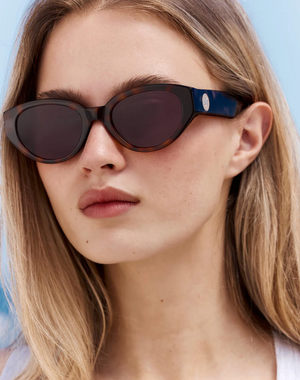 A woman with straight blonde hair and a neutral expression is wearing Jimmy Fairly’s Meli Icons sunglasses, which feature a chic cellulose acetate butterfly frame and dark oval lenses, set against a light blue background.