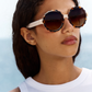 A woman with wavy brown hair wearing the Jimmy Fairly 'Lily' round, scalloped sunglasses with a stainless steel frame and a white sleeveless top stands outdoors against a softly blurred blue background.