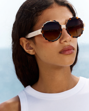 A woman with wavy brown hair wearing the Jimmy Fairly 'Lily' round, scalloped sunglasses with a stainless steel frame and a white sleeveless top stands outdoors against a softly blurred blue background.