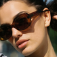 A woman wearing the Jimmy Fairly "Cinnamon" brown oval frame sunglasses with metal hinge accents and a grey shirt looks slightly upward in natural sunlight.