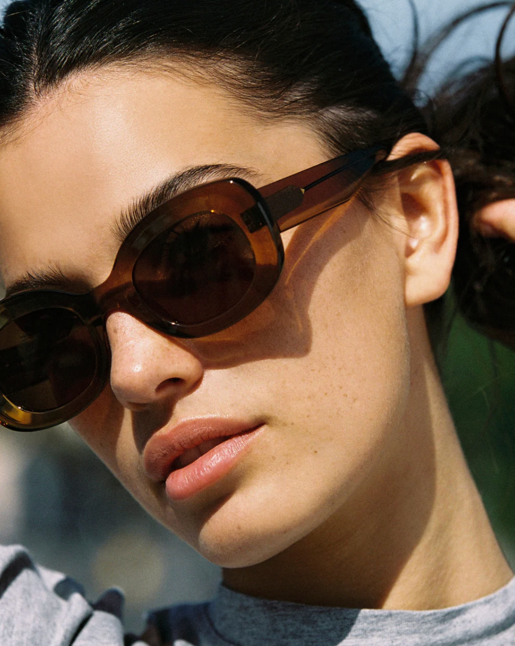 A woman wearing the Jimmy Fairly "Cinnamon" brown oval frame sunglasses with metal hinge accents and a grey shirt looks slightly upward in natural sunlight.