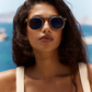 A woman with long dark hair wears the Astie sunglasses by Jimmy Fairly, featuring a sleek stainless steel frame, and a cream-colored top as she stands outdoors against a blurred ocean background.
