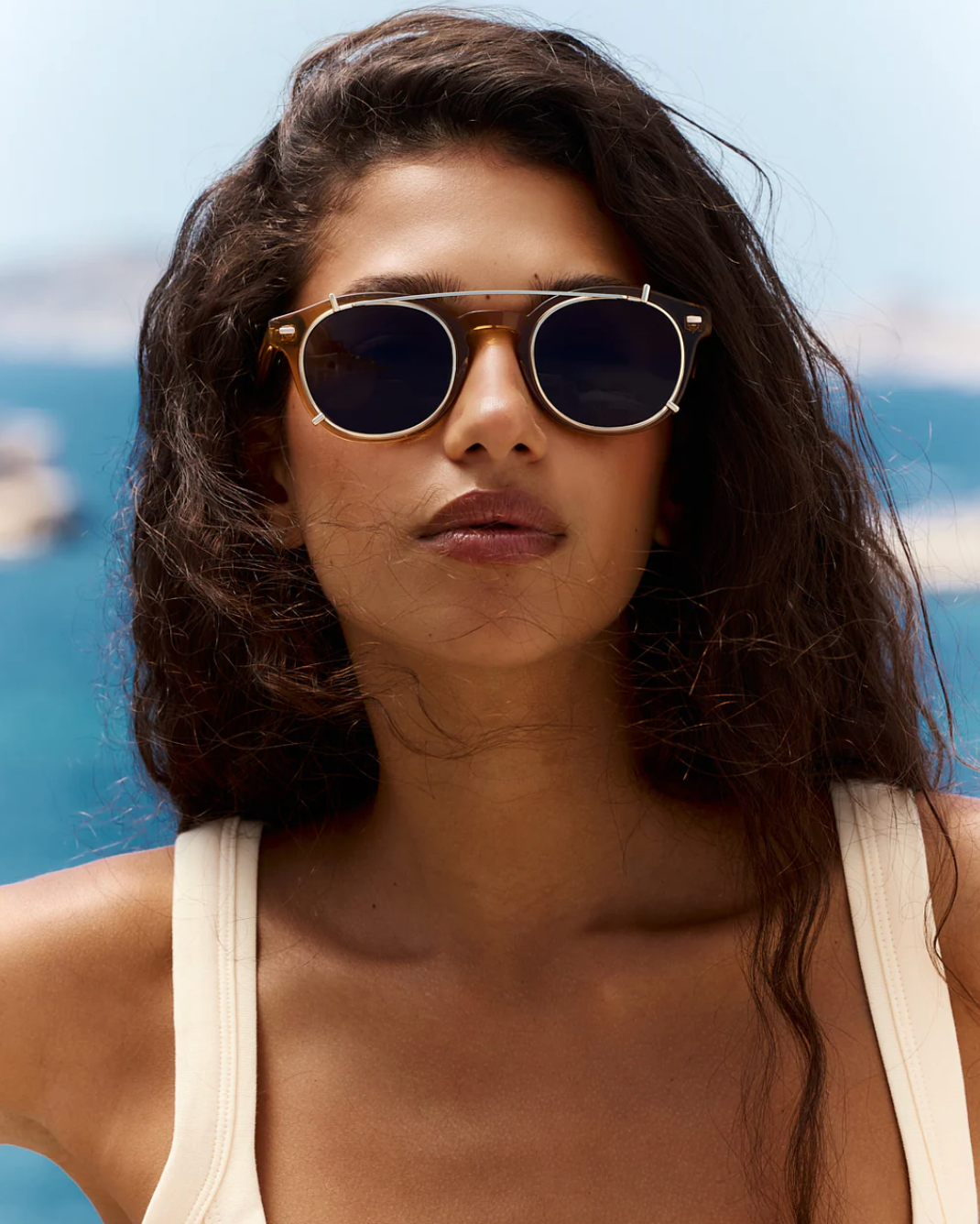 A woman with long dark hair wears the Astie sunglasses by Jimmy Fairly, featuring a sleek stainless steel frame, and a cream-colored top as she stands outdoors against a blurred ocean background.