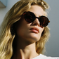 A person with long blonde hair is wearing Jimmy Fairly’s West Coast sunglasses, which feature a cellulose acetate frame and metal hinge, along with a white top while standing outdoors in sunlight.
