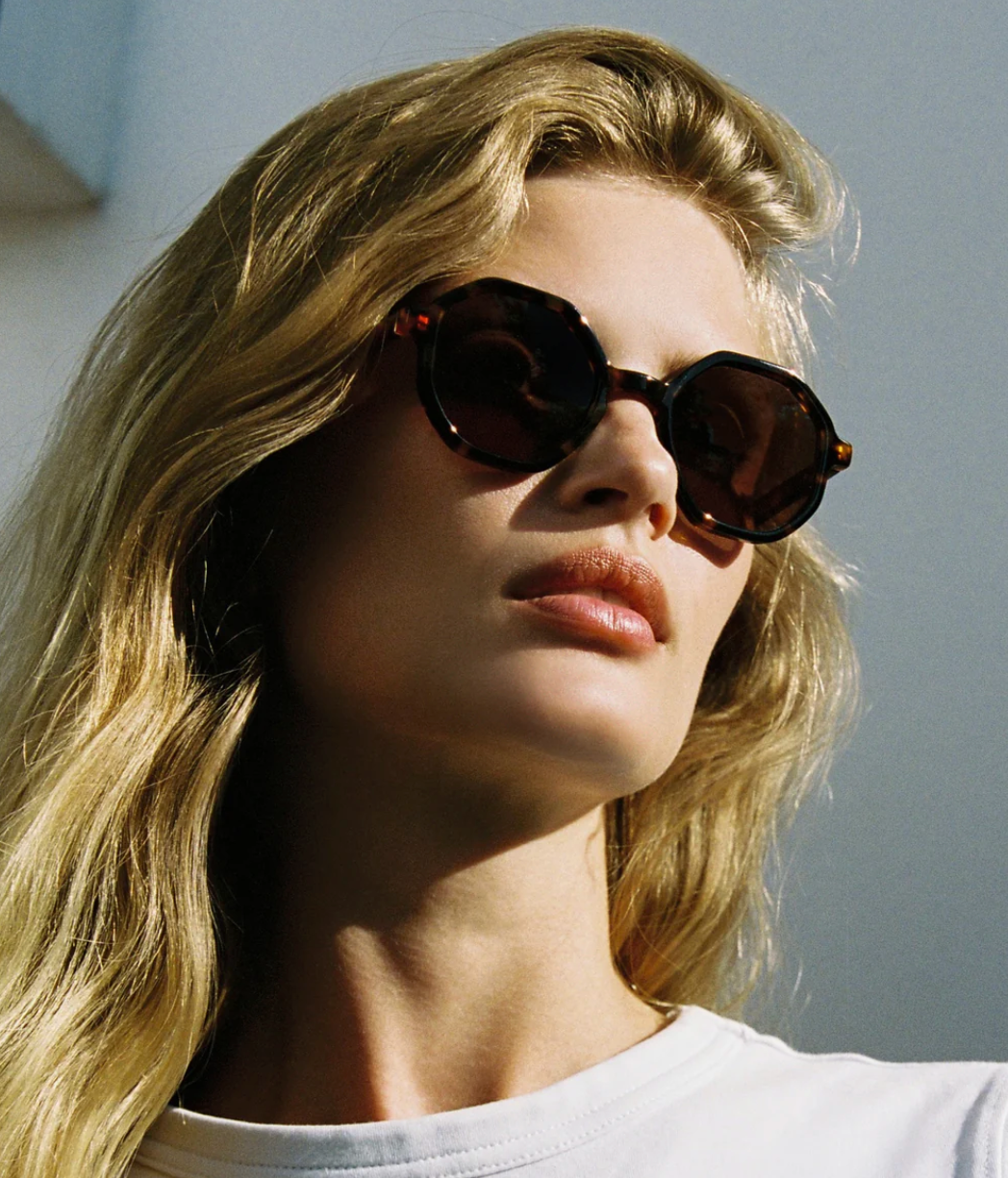 A person with long blonde hair is wearing Jimmy Fairly’s West Coast sunglasses, which feature a cellulose acetate frame and metal hinge, along with a white top while standing outdoors in sunlight.