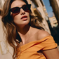 Woman with wavy brown hair wearing the Jimmy Fairly Pippa black sunglasses, which feature a cellulose acetate butterfly frame, and an off-the-shoulder yellow top stands outside on a sunny day, with buildings blurred in the background.
