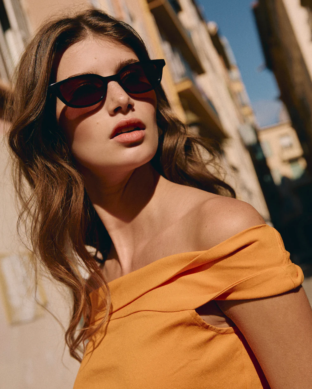 Woman with wavy brown hair wearing the Jimmy Fairly Pippa black sunglasses, which feature a cellulose acetate butterfly frame, and an off-the-shoulder yellow top stands outside on a sunny day, with buildings blurred in the background.