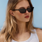 A person with long blonde hair, wearing Jimmy Fairly’s Pippa black butterfly-frame sunglasses and a white tank top, stands outdoors against a clear blue sky.