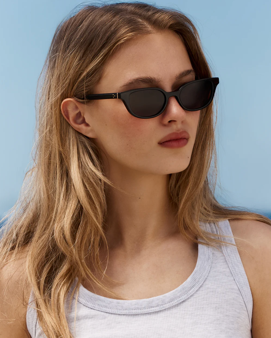 A person with long blonde hair, wearing Jimmy Fairly’s Pippa black butterfly-frame sunglasses and a white tank top, stands outdoors against a clear blue sky.