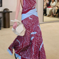 A model walks on a beige carpet wearing the Altuzarra Moonlight Dress, a maroon and light blue patterned silk piece with a high neckline, holding a white bag, while seated people are visible in the background.
