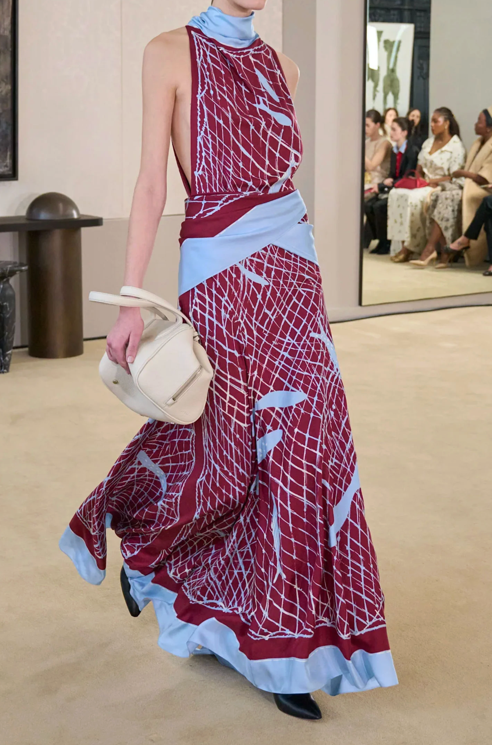 A model walks on a beige carpet wearing the Altuzarra Moonlight Dress, a maroon and light blue patterned silk piece with a high neckline, holding a white bag, while seated people are visible in the background.