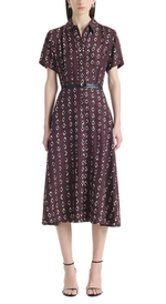 A woman wears the Altuzarra Kiera Dress, a knee-length, short-sleeved maroon pure silk dress featuring a geometric white pattern, cinched at the waist with a leather belt and paired with black strappy heels.