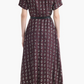 A woman stands facing away, wearing the Altuzarra Kiera Dress—a maroon, short-sleeved midi shirt dress featuring a geometric white pattern and a leather waist belt that accentuates its A-line silhouette—paired with black heels.