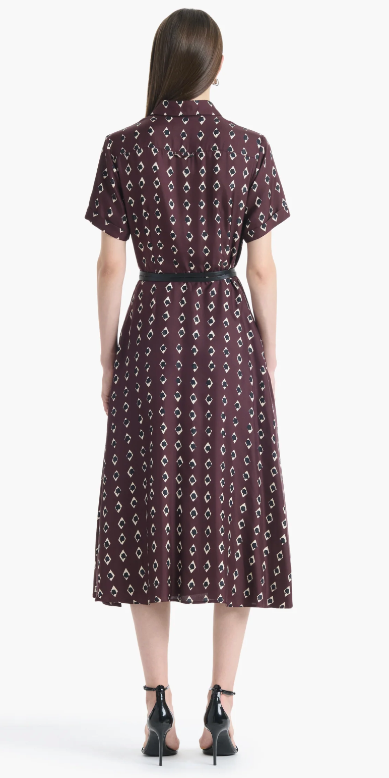 A woman stands facing away, wearing the Altuzarra Kiera Dress—a maroon, short-sleeved midi shirt dress featuring a geometric white pattern and a leather waist belt that accentuates its A-line silhouette—paired with black heels.