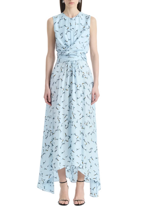 A woman wears the Altuzarra Penny Dress—a sleeveless, light blue silk piece featuring a fitted bodice, bird print, gathered waist, and an asymmetric hem—paired with black strappy heels, standing against a white background.