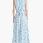 A woman stands with her back to the camera, wearing the Altuzarra Penny Dress—a sleeveless, light blue silk, ankle-length dress adorned with a bird print, featuring an asymmetric hem and a waist tie. She completes the look with black heels.