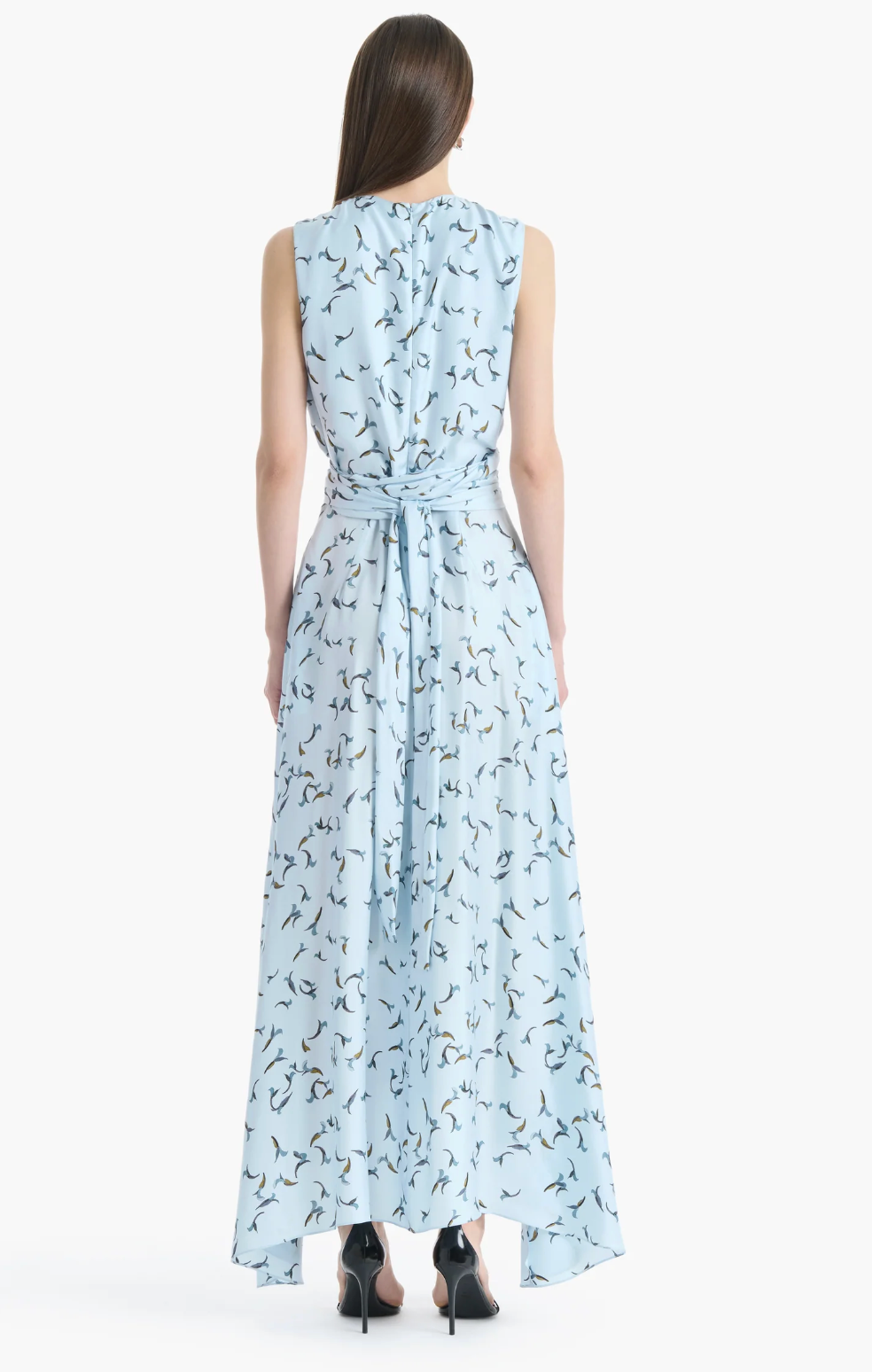 A woman stands with her back to the camera, wearing the Altuzarra Penny Dress—a sleeveless, light blue silk, ankle-length dress adorned with a bird print, featuring an asymmetric hem and a waist tie. She completes the look with black heels.
