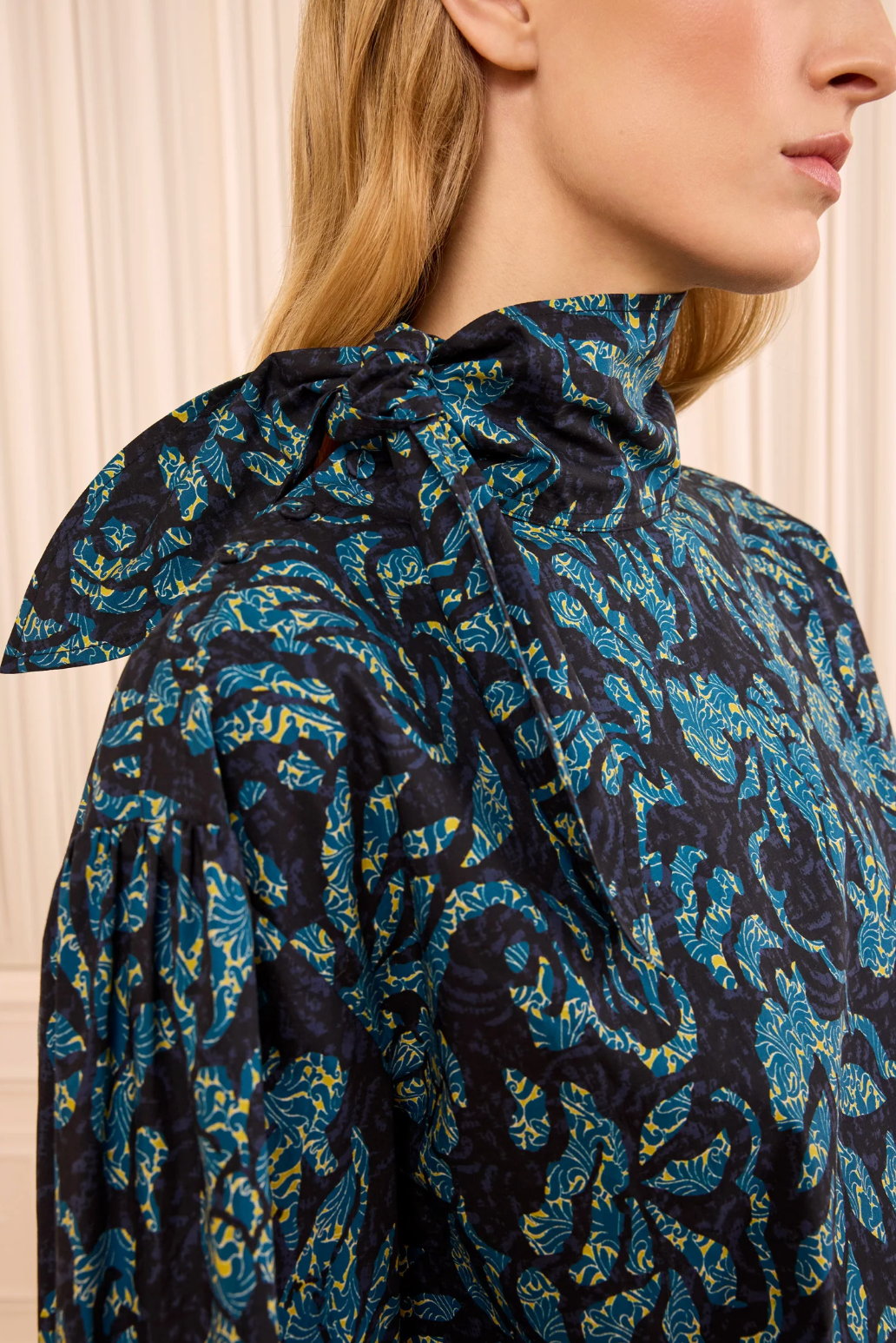 A woman wearing the Ulla Johnson Pastel Long Sleeve Cotton Blouse stands in front of a cream-colored background, featuring a high-neck with a tied bow, long sleeves, and a blue and green floral design that creates an elegant, structured silhouette.