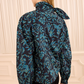 A person with straight, light brown hair is seen from behind, wearing the Ulla Johnson Pastel Long Sleeve Cotton Blouse—featuring a blue and black floral print, tied neck, and structured silhouette—paired with black pants and standing in front of cream-colored drapes.