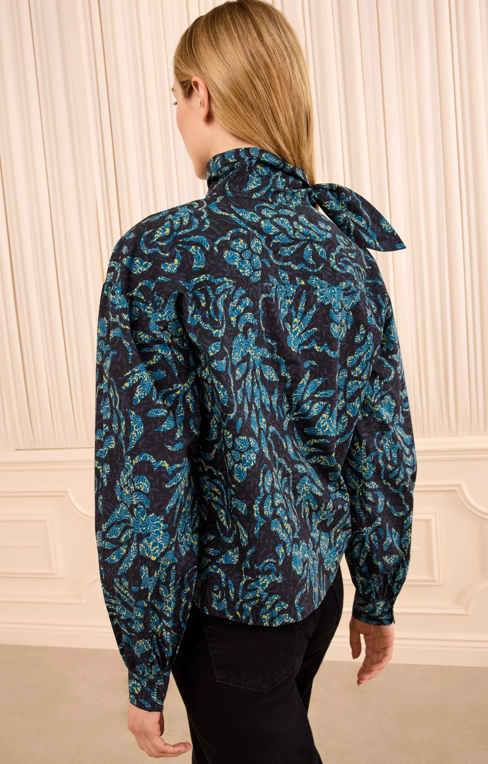 A person with straight, light brown hair is seen from behind, wearing the Ulla Johnson Pastel Long Sleeve Cotton Blouse—featuring a blue and black floral print, tied neck, and structured silhouette—paired with black pants and standing in front of cream-colored drapes.
