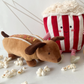 Mon Ami’s Hot Dog - Tan plush, featuring whimsical arms and legs, sits among scattered popcorn pieces on a white surface, accompanied by a playful dachshund toy for extra fun.