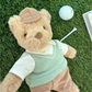 The Mon Ami Bogey Golf Bear, dressed in charming country-club style golf attire, lies on grass next to a golf ball, a white tee, and part of an open book.