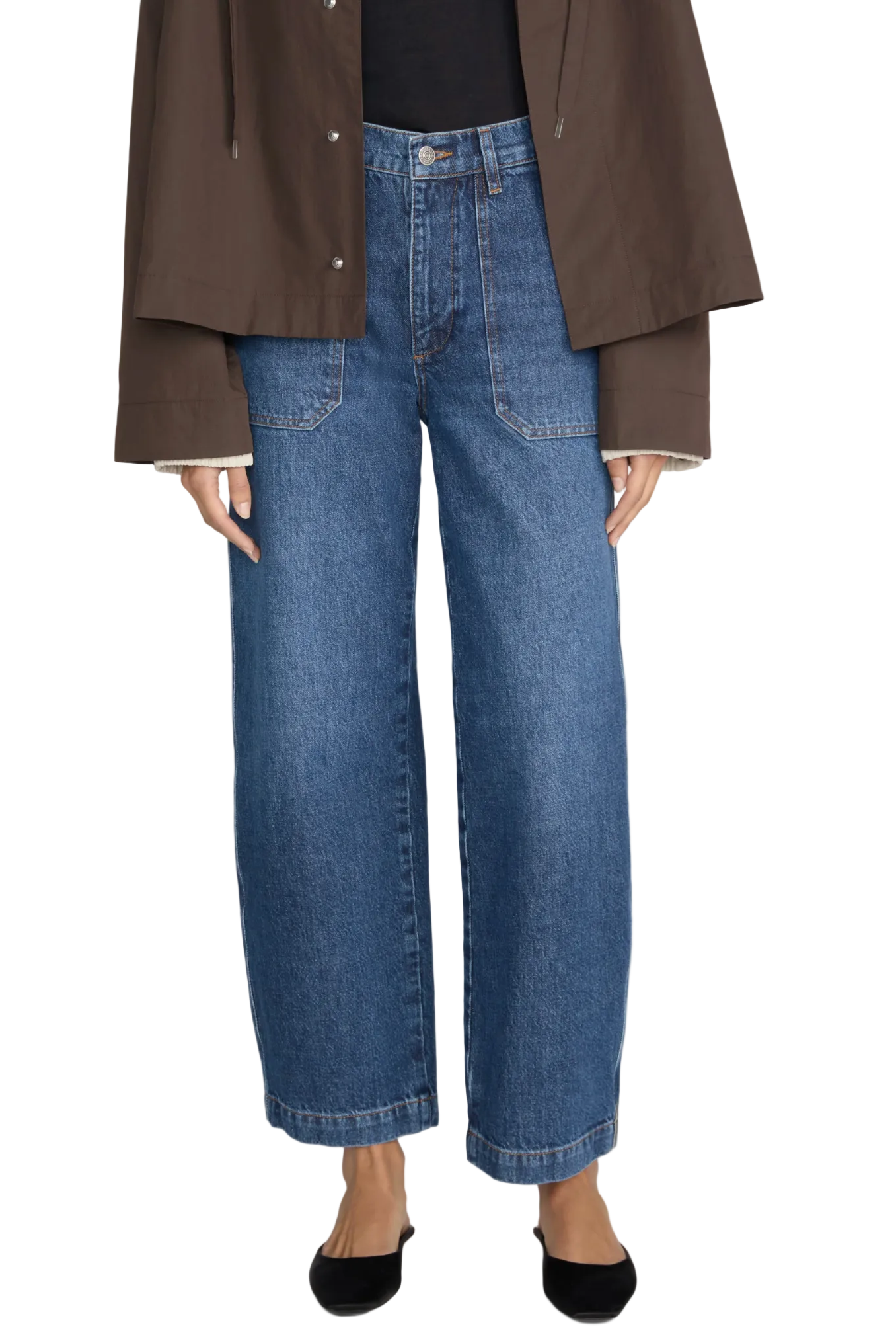 A person wearing Frame’s The Ranger Pant—a pair of high-rise, cropped wide-leg blue jeans crafted from stretch cotton twill with utility front pockets—along with a brown jacket and black flats stands against a plain background.