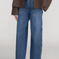 Person wearing Frame’s The Ranger Pant—high-waisted blue wide-leg jeans with utility pocket details—paired with a dark brown jacket and black pointed-toe flats, standing against a plain light background.
