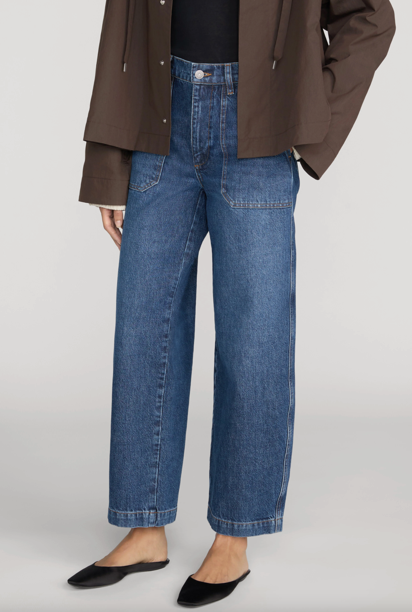 Person wearing Frame’s The Ranger Pant—high-waisted blue wide-leg jeans with utility pocket details—paired with a dark brown jacket and black pointed-toe flats, standing against a plain light background.