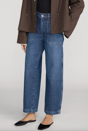 Person wearing Frame’s The Ranger Pant—high-waisted blue wide-leg jeans with utility pocket details—paired with a dark brown jacket and black pointed-toe flats, standing against a plain light background.