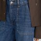 A person wears The Ranger Pant by Frame—high-waisted blue jeans crafted from stretch cotton twill with a silver button—paired with a brown jacket; only the lower torso and part of one hand are visible.